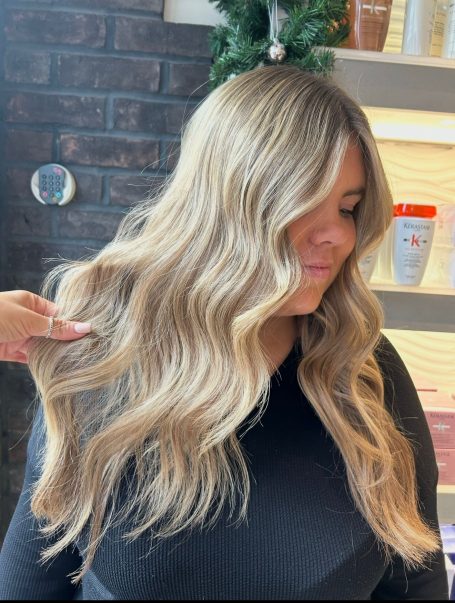 Woman with long, wavy blonde hair, smiling and posing with hand gently touching her hair.